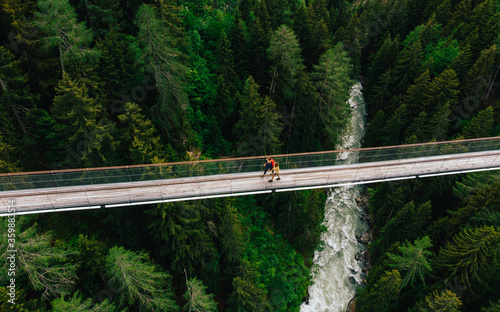 Aerial view of rope walkway through treetops in coniferous forest. Bird's eye view of hanging bridge crossing raging mountains river, suspension bridge with walking people traveller
