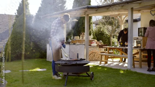 Handsome man cooking preparing food on a bbq fire pit, two people in the background preparing for picnic