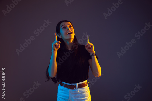 Pointing, choosing. Caucasian young woman's portrait on dark studio background in neon. Beautiful female brunette model in casual. Concept of human emotions, facial expression, sales, ad. Copyspace.