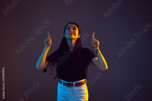 Pointing, choosing. Caucasian young woman's portrait on dark studio background in neon. Beautiful female brunette model in casual. Concept of human emotions, facial expression, sales, ad. Copyspace.