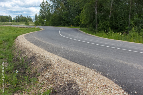 View to a winding new asphalt road in a rural area. Gravel roadside, ditch for rainwater. Latvia. 