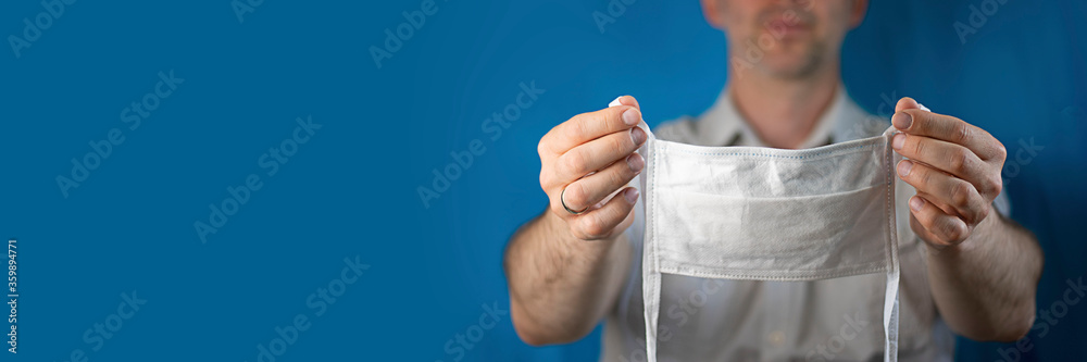 Preventive white medical mask in man's hands on the background of defocused unrecognizable man wearing shirt, holds out arms. Indoor studio shot on blue background, copy space. Horizontal banner