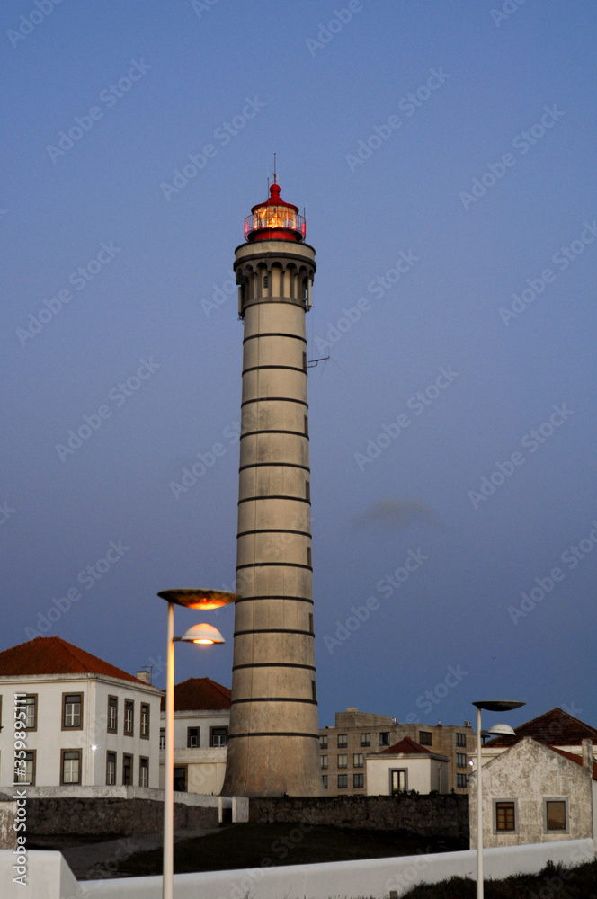 High lighthouse on the background of Purple sky, luminous lantern. Houses near Lighthouse