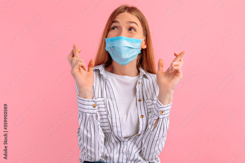Focused girl in a medical protective mask on her face, fingers crossed for good luck, on a pink background Focused girl in a medical protective mask on her face, fingers crossed for good luck, on a pink background