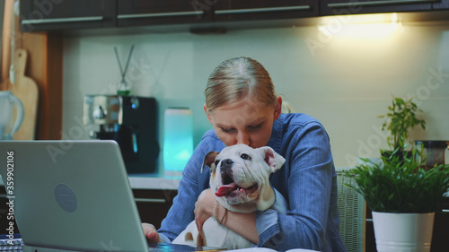 Close-up shot of cheerful woman hugging small dog in the kitchen. She working on the computer and holding small dog on her hands