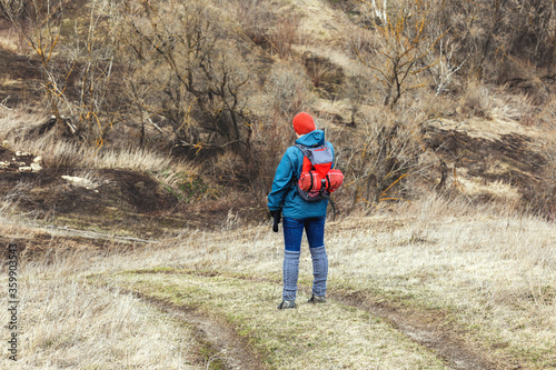 A tourist girl in a sports jacket and hat is walking along a dirt road on a sunny spring day. Hiking, healthy lifestyle. © Светлана Горбань