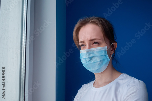 Portrait of pensive woman wearing medical face mask and looking out of window in room with blue wall at home. Self isolation, prevention, quarantine, COVID-19, coronavirus, safety concept