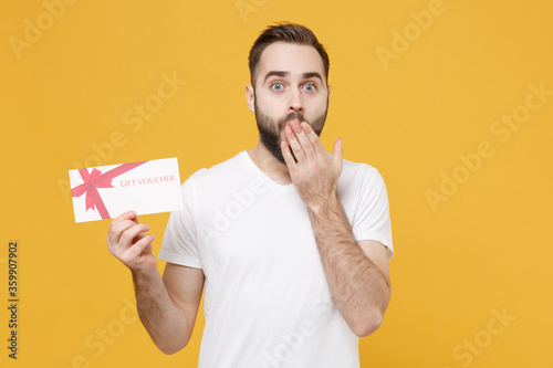 Shocked young bearded man guy in white casual t-shirt posing isolated on yellow wall background studio. People lifestyle concept. Mock up copy space. Hold gift certificate, covering mouth with hand. © ViDi Studio