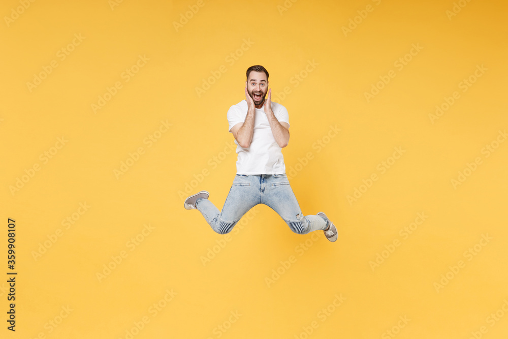 Surprised young bearded man guy in white casual t-shirt posing isolated on yellow background studio portrait. People lifestyle concept. Mock up copy space. Jumping spreading legs put hands on cheeks.