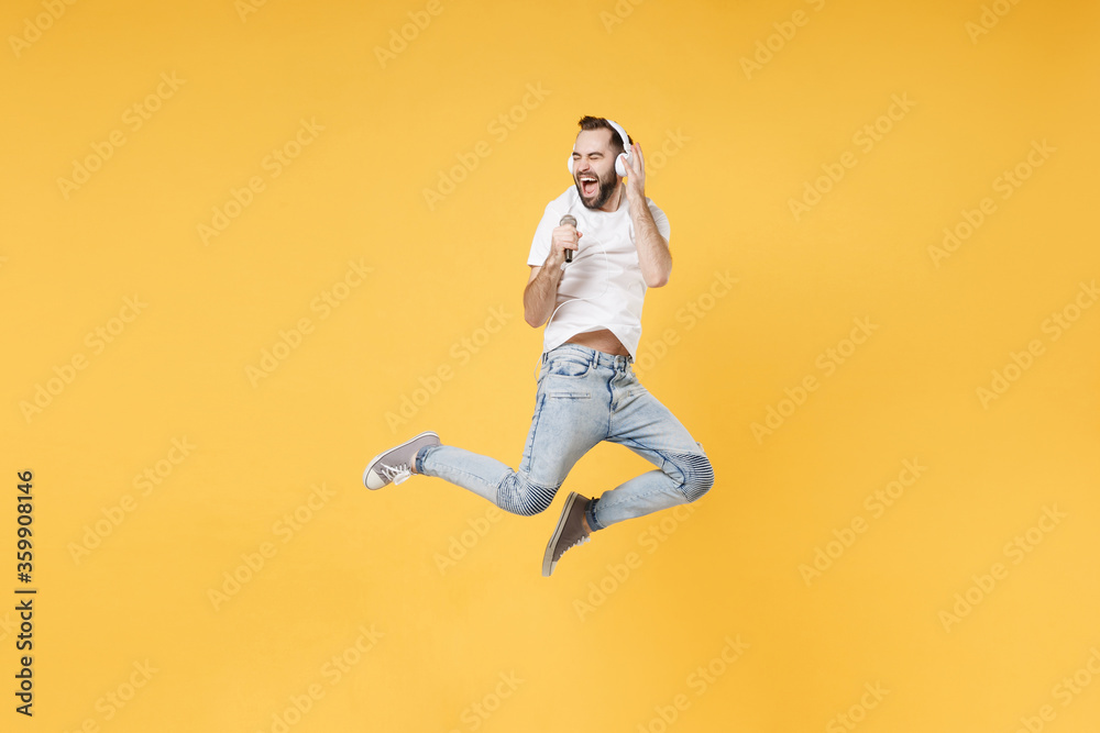 Funny young bearded man guy in white casual t-shirt posing isolated on yellow background. People lifestyle concept. Mock up copy space. Jumping, listen music with headphones, sing song in microphone.
