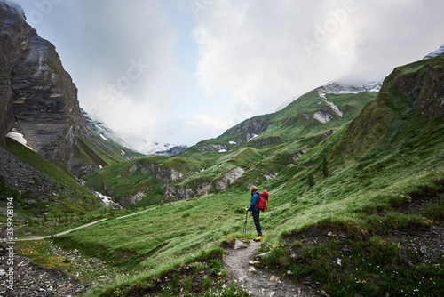 Man tourist with backpack standing on rocky mountain path in green hillside valley. Mountaineer enjoying the view of beautiful high rocks and cliffs while traveling alone. Concept of hiking, traveling