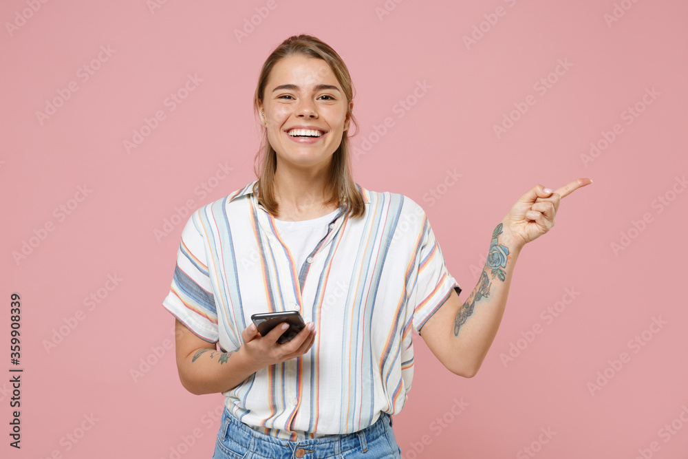 Smiling young blonde woman girl in casual striped shirt posing isolated on pastel pink background studio. People lifestyle concept. Mock up copy space. Using mobile phone pointing index finger aside. Smiling young blonde woman girl in casual striped shirt posing isolated on pastel pink background studio. People lifestyle concept. Mock up copy space. Using mobile phone pointing index finger aside.