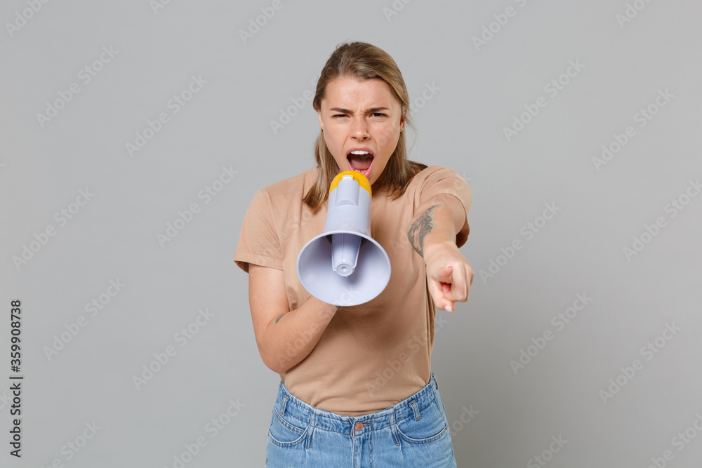 Angry young blonde woman girl in casual beige t-shirt posing isolated on gray background studio. People lifestyle concept. Mock up copy space. Screaming in megaphone, pointing index finger on camera. Angry young blonde woman girl in casual beige t-shirt posing isolated on gray background studio. People lifestyle concept. Mock up copy space. Screaming in megaphone, pointing index finger on camera.