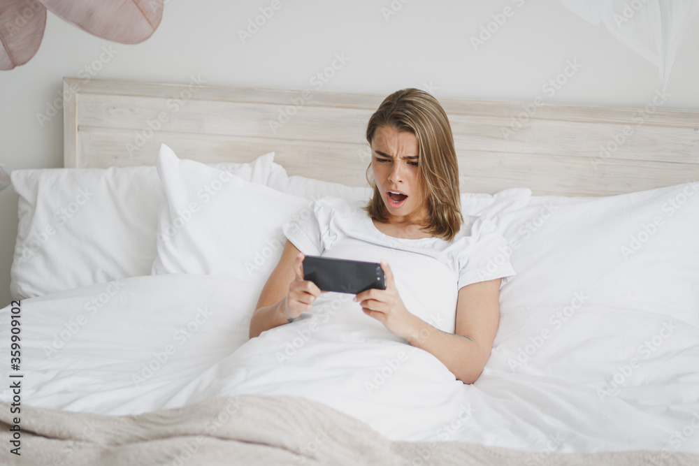 Displeased girl lying in bed with white sheet pillow blanket spending time in bedroom at home. Rest relax good mood lifestyle concept. Mock up copy space. Using mobile phone, play game watching video. Displeased girl lying in bed with white sheet pillow blanket spending time in bedroom at home. Rest relax good mood lifestyle concept. Mock up copy space. Using mobile phone, play game watching video.