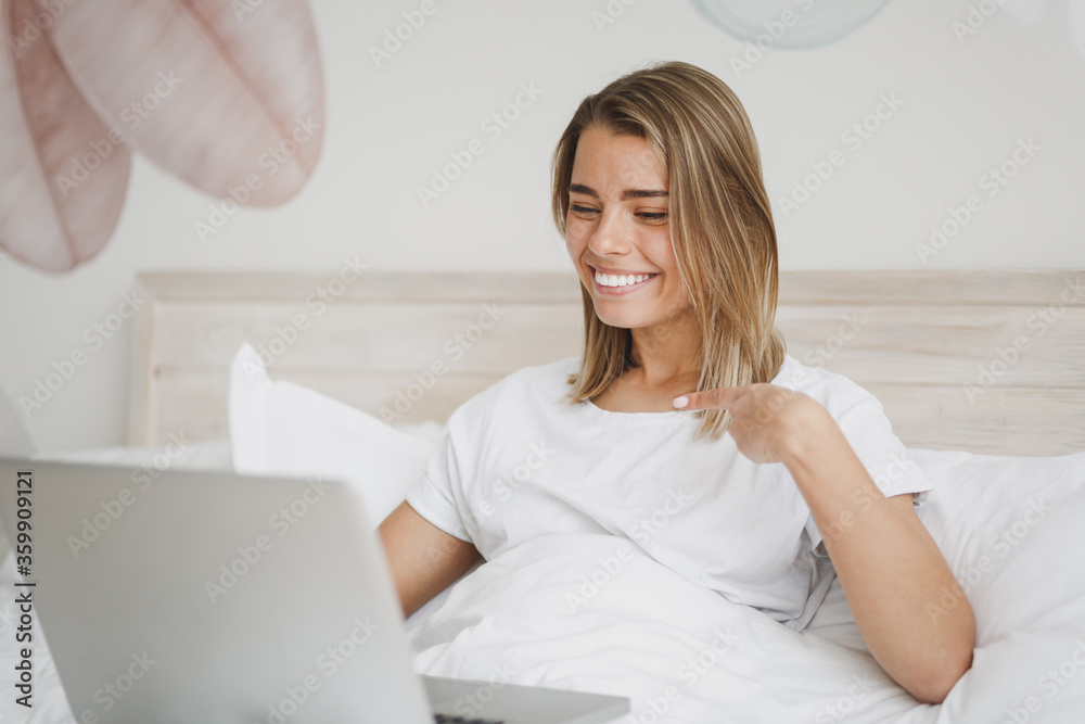 Smiling girl lying in bed with white sheet pillow blanket spending time in bedroom at home. Rest relax good mood lifestyle concept. Mock up copy space. Working on laptop pc computer point on herself. Smiling girl lying in bed with white sheet pillow blanket spending time in bedroom at home. Rest relax good mood lifestyle concept. Mock up copy space. Working on laptop pc computer point on herself.