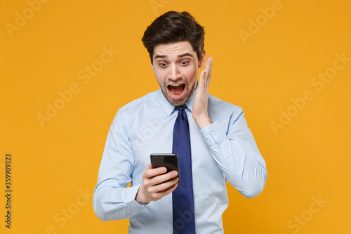 Shocked young business man in classic blue shirt tie posing isolated on yellow background. Achievement career wealth business concept. Mock up copy space. Using mobile phone scream put hand on cheek.