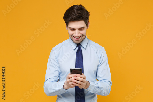 Smiling young business man in classic blue shirt tie posing isolated on yellow background studio. Achievement career wealth business concept. Mock up copy space. Using mobile phone typing sms message.