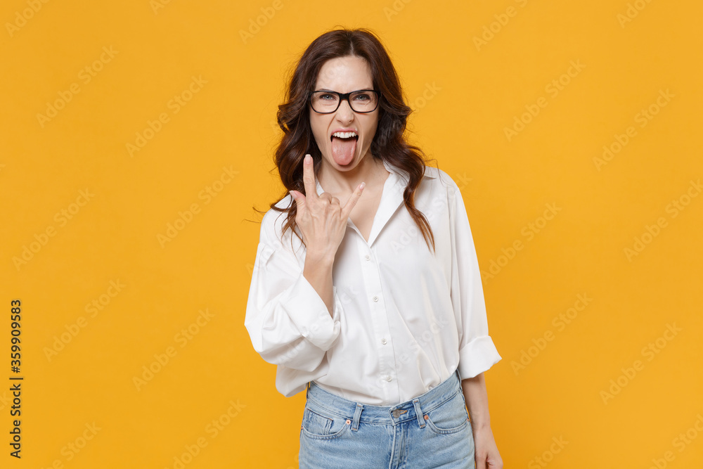 Crazy young brunette business woman in white shirt glasses isolated on yellow background. Achievement career wealth business concept. Depicting heavy metal rock sign, horns up gesture, showing tongue.