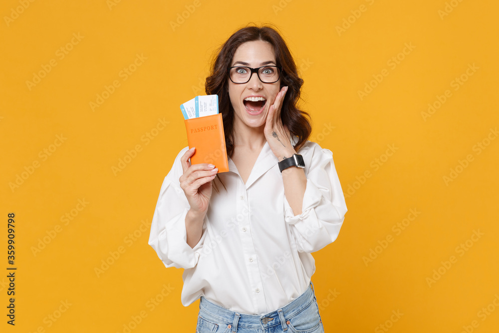 Surprised young brunette business woman in white shirt glasses isolated on yellow background. Achievement career wealth business concept. Mock up copy space. Hold passport tickets, put hand on cheek.