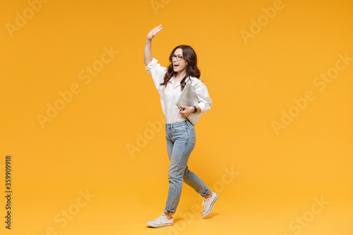 Excited young business woman in white shirt glasses isolated on yellow background. Achievement career wealth business concept. Hold laptop pc computer waving and greeting with hand as notices someone. © ViDi Studio