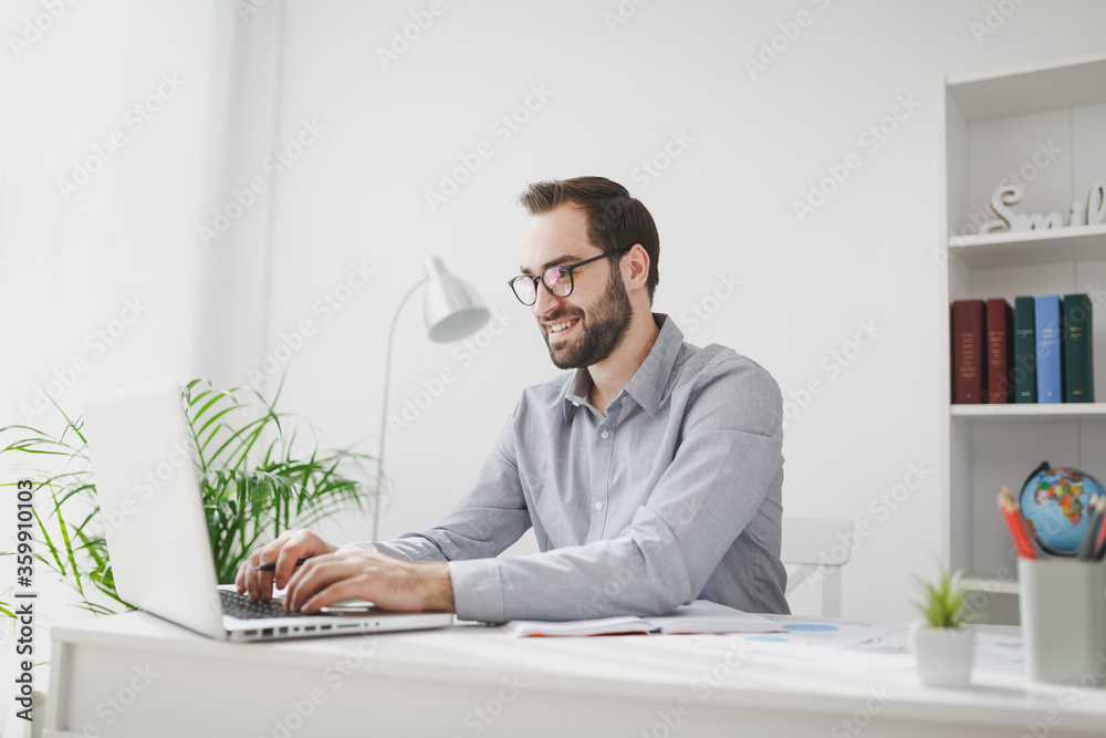 Smiling young bearded business man in gray shirt glasses sitting at desk in light office on white wall background. Achievement business career concept. Working on laptop pc computer, holding pen.