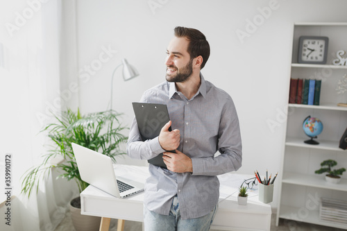 Smiling bearded business man in gray shirt stand near desk with laptop pc computer in light office on white wall background. Achievement business career concept. Hold clipboard with papers document.