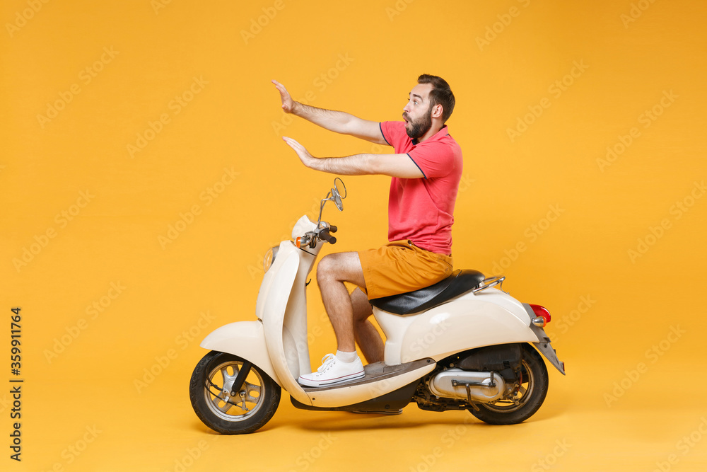 Side view of shocked young bearded man guy in summer clothes driving moped isolated on yellow background. Driving motorbike transportation concept. Mock up copy space. Showing stop gesture with palms. Side view of shocked young bearded man guy in summer clothes driving moped isolated on yellow background. Driving motorbike transportation concept. Mock up copy space. Showing stop gesture with palms.