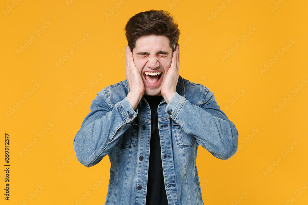 Crazy young man guy in casual denim jacket posing isolated on yellow wall background studio portrait. People lifestyle concept. Mock up copy space. Keeping eyes closed, put hands on cheeks, screaming. Crazy young man guy in casual denim jacket posing isolated on yellow wall background studio portrait. People lifestyle concept. Mock up copy space. Keeping eyes closed, put hands on cheeks, screaming.