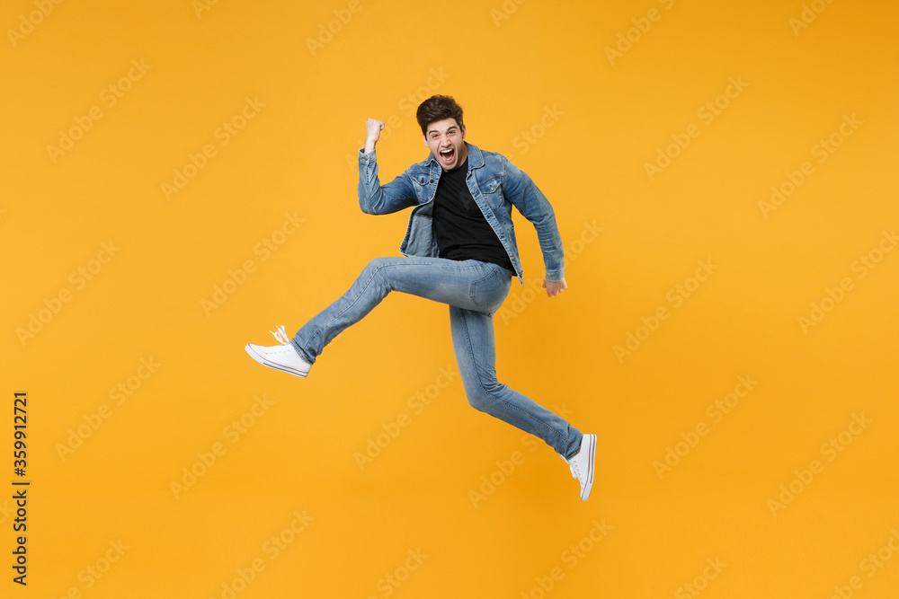 Screaming young man guy wearing casual denim clothes posing isolated on yellow background studio portrait. People lifestyle concept. Mock up copy space. Jumping, spreading legs doing winner gesture.