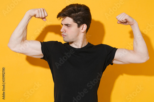 Strong young man guy 20s wearing casual black t-shirt posing isolated on yellow wall background studio portrait. People sincere emotions lifestyle concept. Mock up copy space. Showing biceps, muscles.