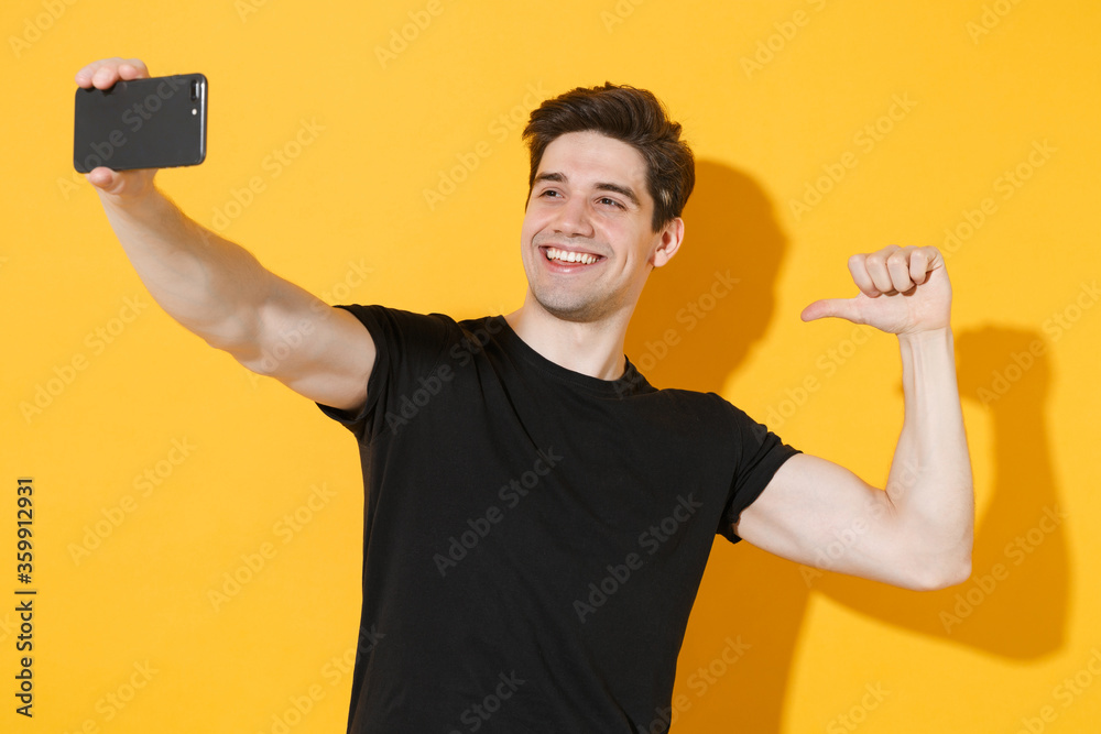 Smiling young man guy in casual black t-shirt posing isolated on yellow background studio. People lifestyle concept. Mock up copy space. Doing selfie shot on mobile phone, pointing thumb on himself. Smiling young man guy in casual black t-shirt posing isolated on yellow background studio. People lifestyle concept. Mock up copy space. Doing selfie shot on mobile phone, pointing thumb on himself.