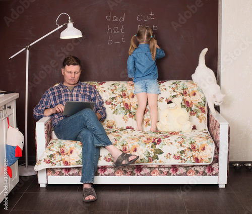 Little girl wrighting on the chalkboard doing English with her cat while her father working on his laptop sitting on the couch. Girl writing missing letters in English words. Home education concept.