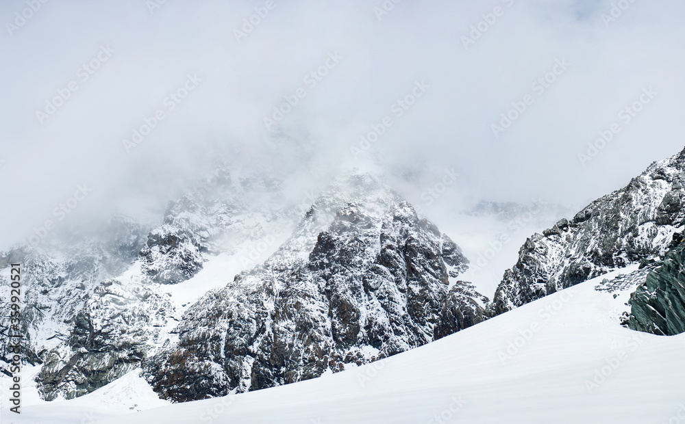 Fantastic scenery of rocky mountains covered with snow. Panoramic view of snow-capped alpine rocks with pyramid-shaped peak under foggy sky. Concept of rocky hills, nature beauty and wintertime.