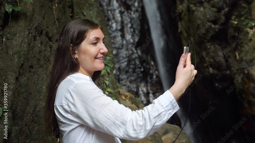 concentrated dark haired girl blogger wearing headphones holds smartphone in hand and conducts video call against tropical waterfall close view concentrated dark haired girl blogger wearing headphones holds smartphone in hand and conducts video call against tropical waterfall close view