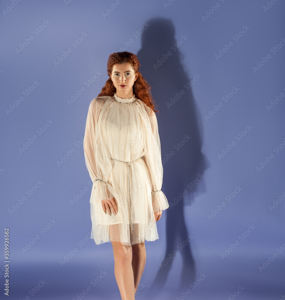 Isolated slender red-haired curly-haired girl in a white designer dress on a blue background. Wedding elegant dress.
