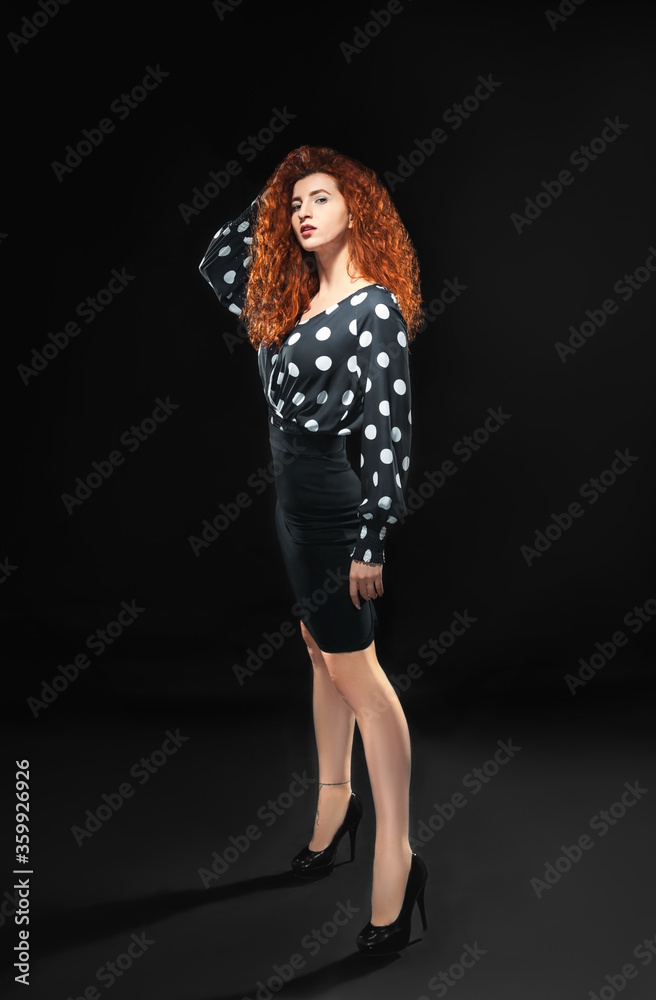 Isolated slender red-haired curly girl in a blouse and a black skirt against a black background. Office employee. Isolated slender red-haired curly girl in a blouse and a black skirt against a black background. Office employee.