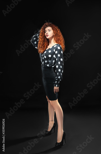 Isolated slender red-haired curly girl in a blouse and a black skirt against a black background. Office employee.