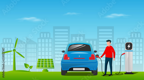 Electric car charging at the charger station with a young man holding the cable. Wind turbine and solar panel with green city skyline in background