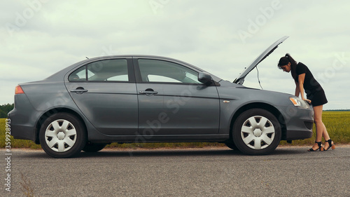 A young woman near a wrecked car on the side of the road looks under the hood, trying to fix the car.