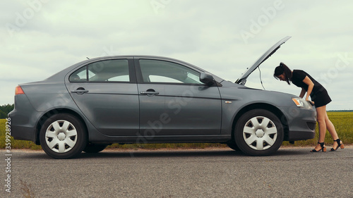 A young woman near a wrecked car on the side of the road looks under the hood, trying to fix the car.