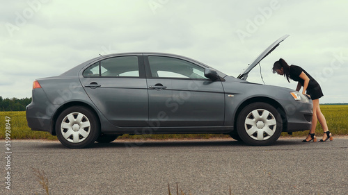 A young girl in a short dress approaches a broken car and opens the hood.The car suddenly broke down on a country road down while driving.