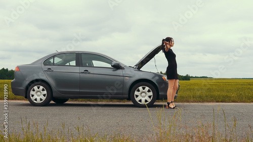 A young girl in a short dress near a broken car is trying to stop a car. The car suddenly broke down on a country road down while driving.		
