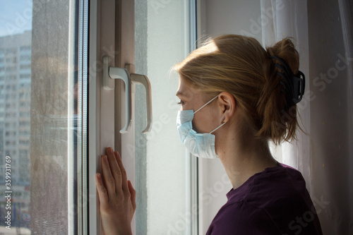 young blonde girl in a white blouse and a medical mask sits outside window and looks outside. Quarantine and home isolation from coronavirus.