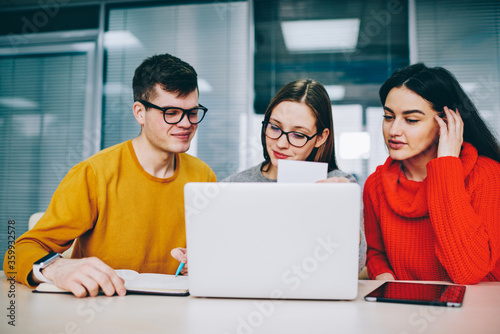 Successful hipster student rewriting information from business card in search job.Positive young people dressed in casual wear collaborating at laptop computer in office sitting at modern laptop