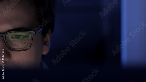 Man in eyeglasses late at night scrolling in front of laptop half of the screen. Coder, programmer or developer using laptop in dark. Close up of glasses with reflection of computer screen.