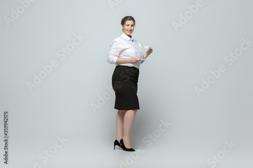 Coffee time. Young woman in office attire. Bodypositive female character, feminism, loving herself, beauty concept. Plus size businesswoman on gray background. Boss, beautiful. Inclusion, diversity.