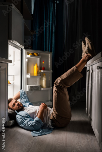 Side view of man looking at camera while lying on floor near open fridge at night