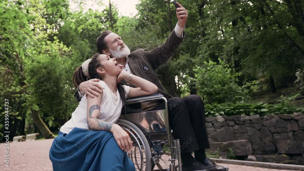 Family concept where happy smiling attractive modern granddaughter with dreadlocks and retired bearded granddad in wheelchair making selfie in urban park,4k Family concept where happy smiling attractive modern granddaughter with dreadlocks and retired bearded granddad in wheelchair making selfie in urban park,4k