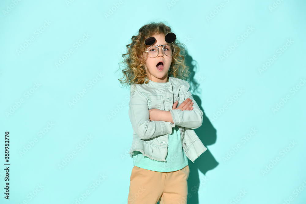 Lovely blond girl with curly hair posing in stylish flip-up glasses calling someone holding hands crossed on chest, isolated on turquoise Lovely blond girl with curly hair posing in stylish flip-up glasses calling someone holding hands crossed on chest, isolated on turquoise