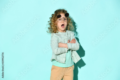 Lovely blond girl with curly hair posing in stylish flip-up glasses calling someone holding hands crossed on chest, isolated on turquoise